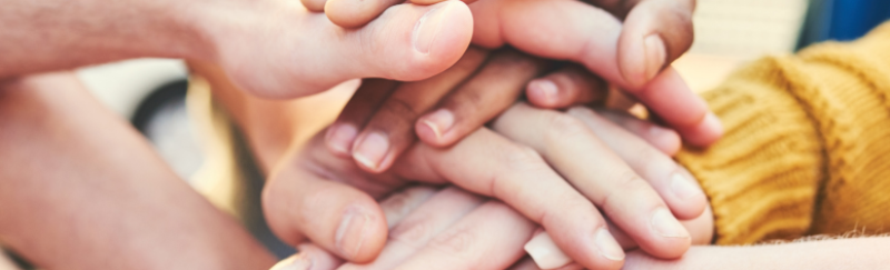 The image shows multiple hands stacked on top of each other, symbolizing unity and teamwork. The hands vary in size, suggesting people of different ages or backgrounds coming together, possibly for a common cause or group effort. The warmth of the image,