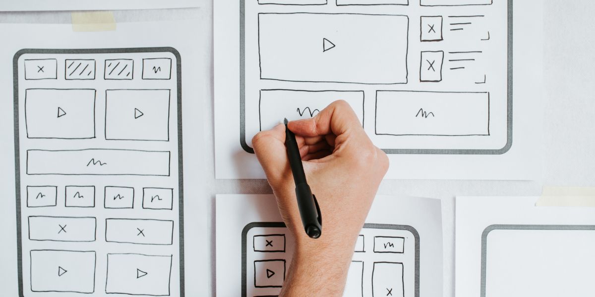 Hand sketching website wireframes with marker on paper, featuring layout boxes, icons, and interface elements for digital design planning.