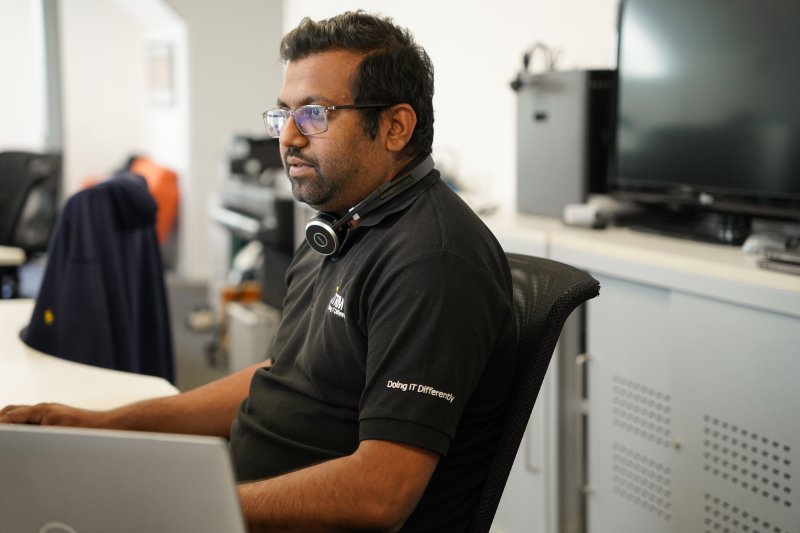 Man with black ITRM branded polo with the logo on the breast and the 'Doing IT Differently' slogan embroidered on the sleeve, sat at his desk looking deep in thought at a computer screen 