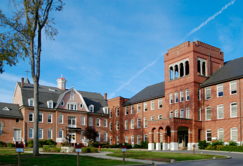 A large, historic brick school building with multiple wings, a central tower, and a cupola, surrounded by trees and manicured lawns under a clear sky with a visible contrail.