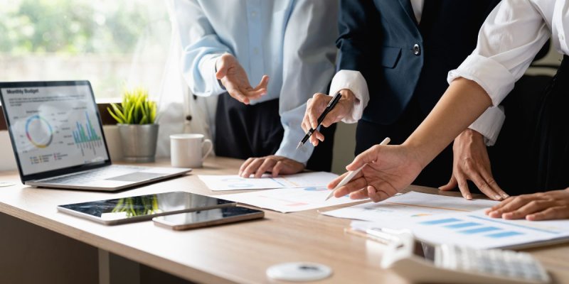 Business professionals in formal attire gathered around a table reviewing documents and charts, with a laptop displaying a 'Monthly Budget' report, surrounded by office items like a tablet, smartphone, coffee cup, and plant.