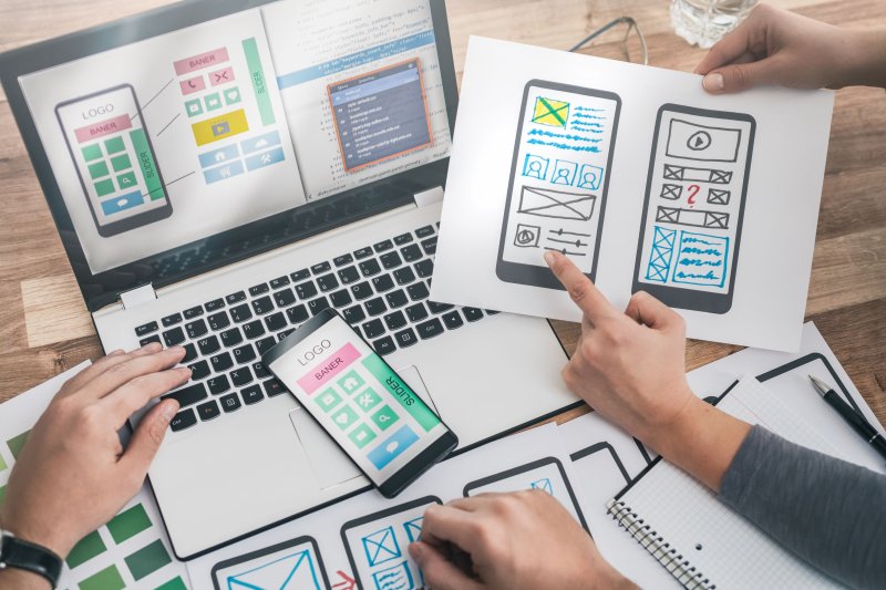 Team collaborating on mobile app design, with laptop mockups, smartphone prototypes, and printed wireframes spread across a workspace.