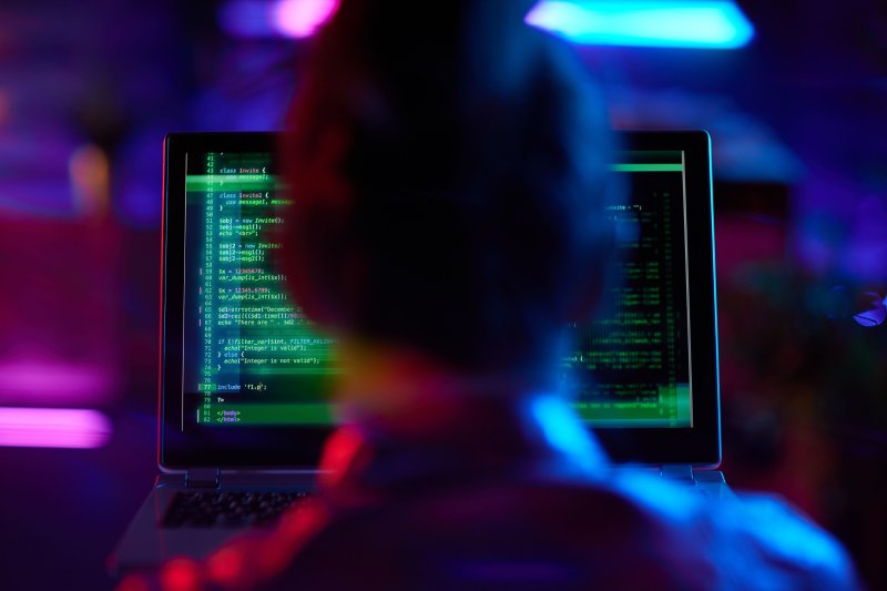 Person working on a laptop displaying green code, with a blurred background of neon blue and pink lights, symbolising digital technology and cybersecurity.